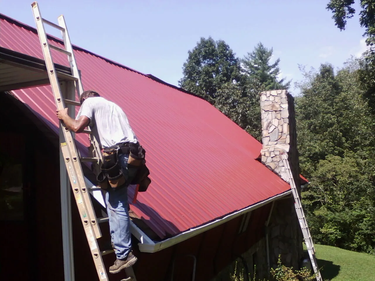 Roofer on a ladder beside a red metal roof and stone chimney — Soffit & Fascia Repair by Roofing Brothers in Belle